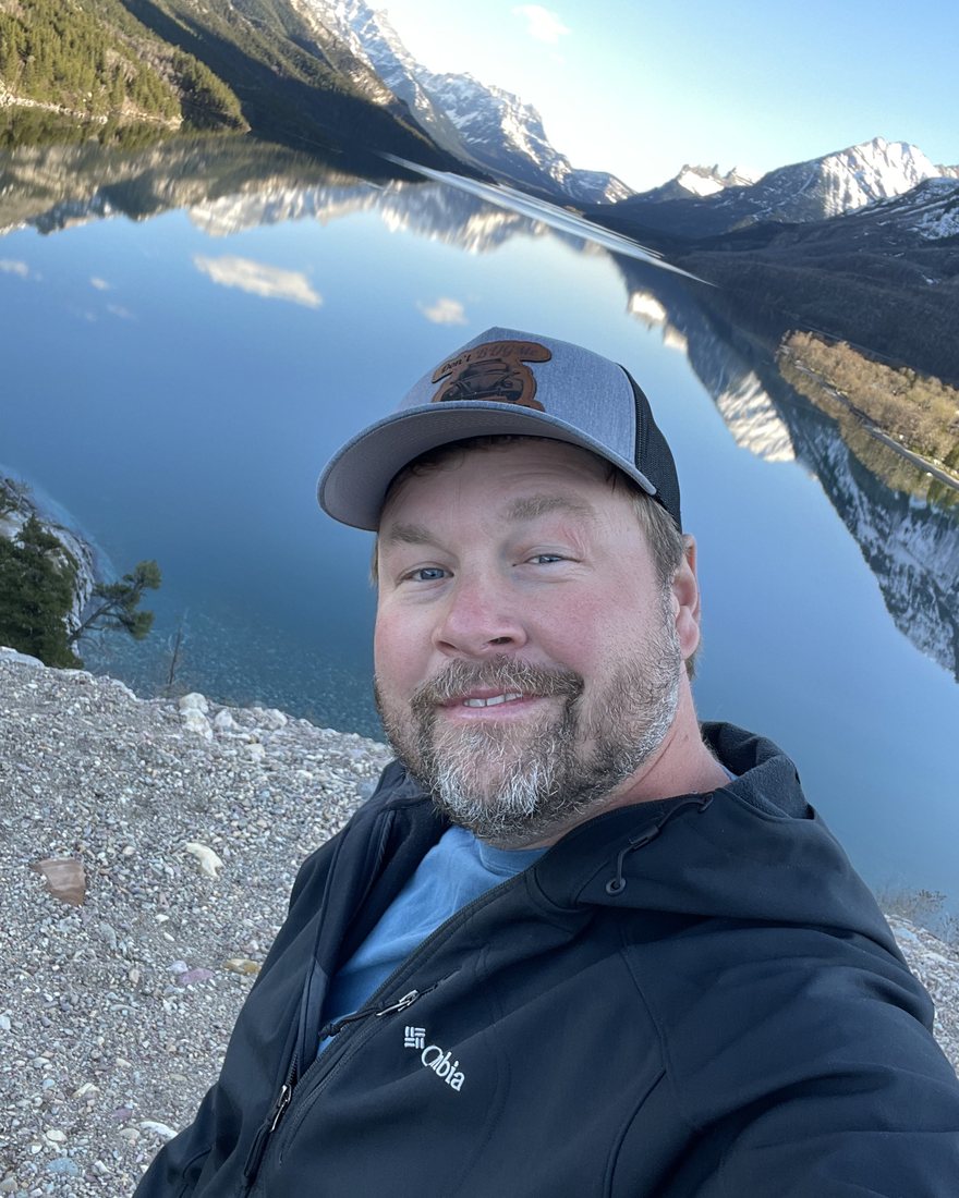 Josh Melcher outdoors above a still mountain lake, wearing a Don't BUGMe cap.